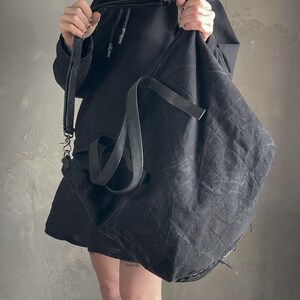 May include: A large, slouchy black shoulder bag made of a textured, distressed fabric. The bag features a long, adjustable black strap with metal hardware clips and a secondary shorter handle. The material has a wrinkled, matte finish throughout.