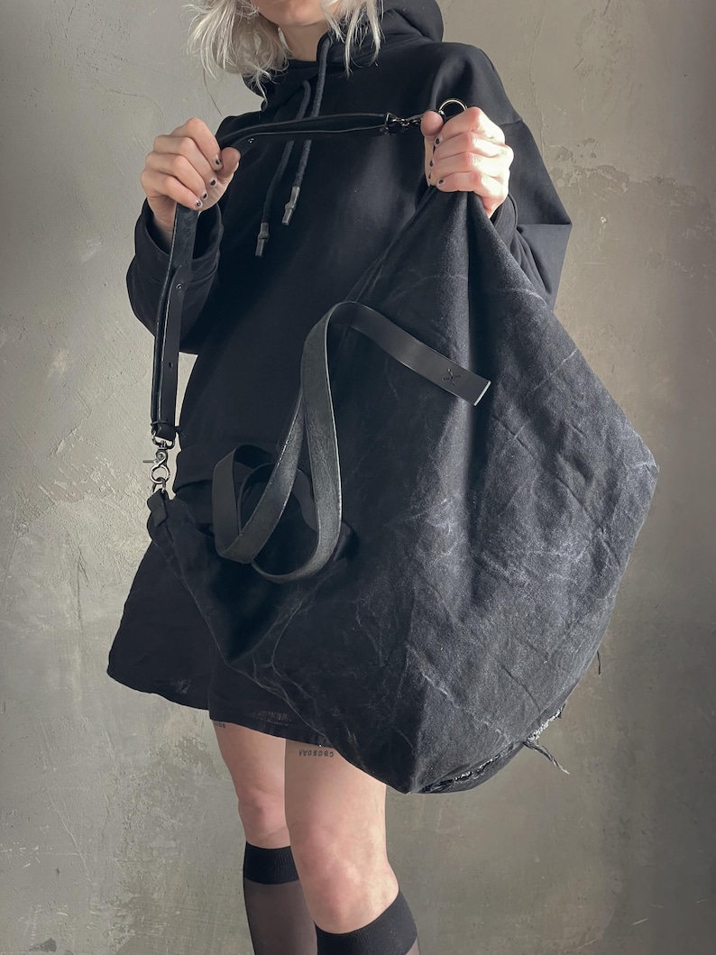 May include: A large, slouchy black shoulder bag made of a textured, distressed fabric. The bag features a long, adjustable black strap with metal hardware clips and a secondary shorter handle. The material has a wrinkled, matte finish throughout.
