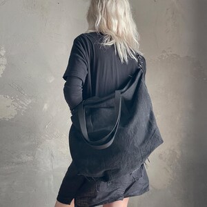 May include: A large, unstructured black tote bag made of a textured fabric. The bag features two wide shoulder straps and a spacious main compartment. It is worn over the shoulder, resting against the side, showcasing a simple and functional design.