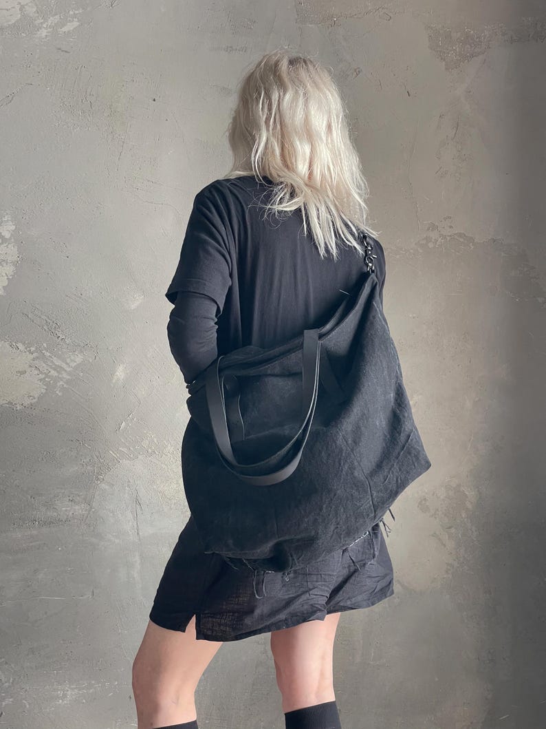 May include: A large, unstructured black tote bag made of a textured fabric. The bag features two wide shoulder straps and a spacious main compartment. It is worn over the shoulder, resting against the side, showcasing a simple and functional design.