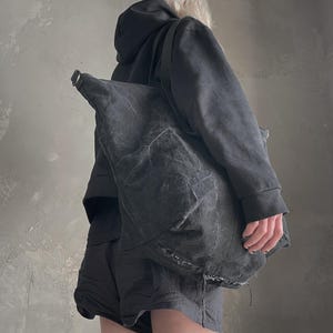 May include: A large, slouchy charcoal grey shoulder bag crafted from a textured, distressed fabric. The bag features a wide strap, a metallic D-ring attachment point, and visible frayed edges along the bottom seam, creating a worn aesthetic.