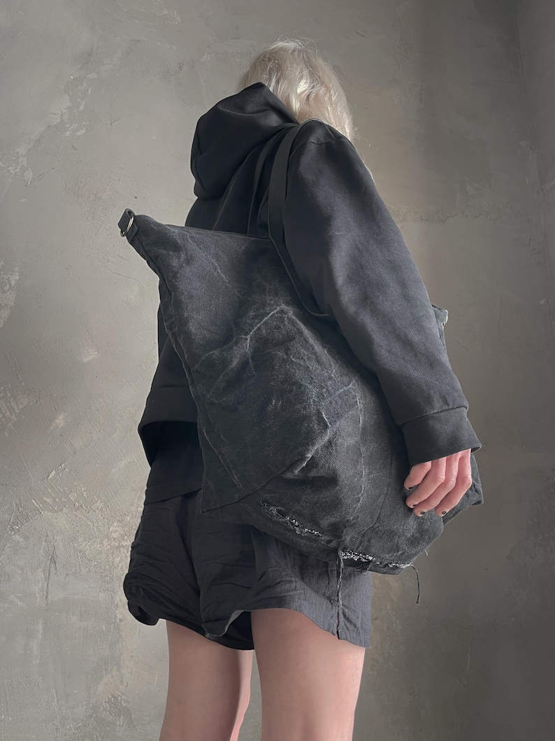 May include: A large, slouchy charcoal grey shoulder bag crafted from a textured, distressed fabric. The bag features a wide strap, a metallic D-ring attachment point, and visible frayed edges along the bottom seam, creating a worn aesthetic.