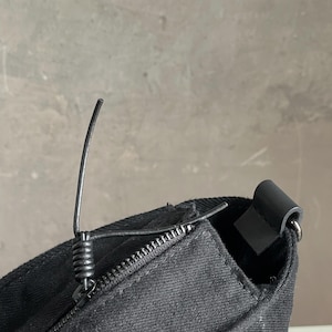 May include: A black canvas bag featuring a top zip closure with a coiled metal pull tab. The bag has a structured seam along the top edge and a dark attachment loop on the side for a strap, showcasing a durable fabric texture throughout.