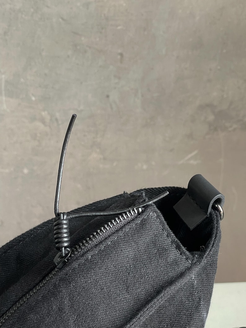 May include: A black canvas bag featuring a top zip closure with a coiled metal pull tab. The bag has a structured seam along the top edge and a dark attachment loop on the side for a strap, showcasing a durable fabric texture throughout.