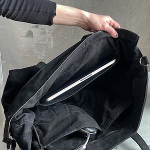 May include: A black canvas tote bag with a zipped top closure, featuring a spacious interior compartment containing a silver laptop and a pair of black over-ear headphones tucked into an internal pocket, with sturdy black fabric shoulder straps.