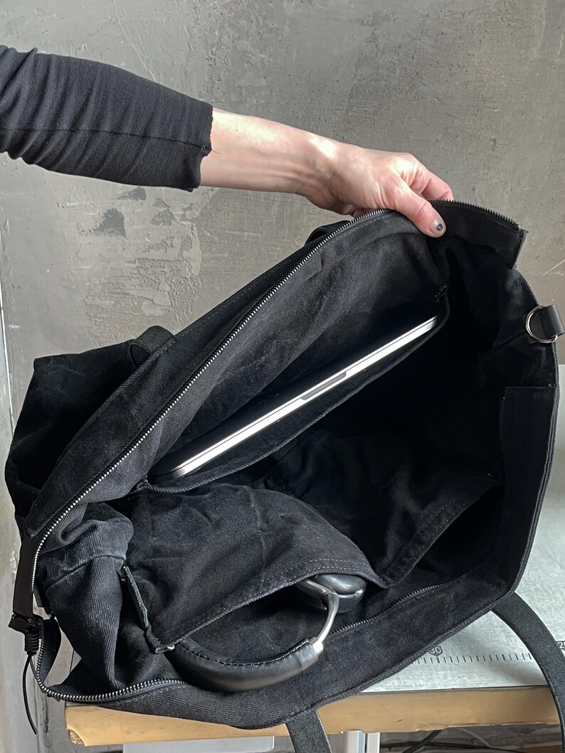 May include: A black canvas tote bag with a zipped top closure, featuring a spacious interior compartment containing a silver laptop and a pair of black over-ear headphones tucked into an internal pocket, with sturdy black fabric shoulder straps.