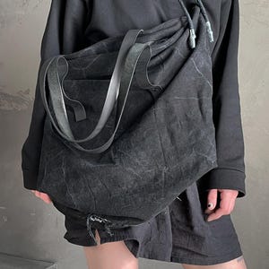 May include: A large, slouchy black shoulder bag made of a textured, distressed fabric. The bag features a wide, flat strap and a drawstring closure at the top with cord ends. The material has a weathered, matte appearance with visible creases.