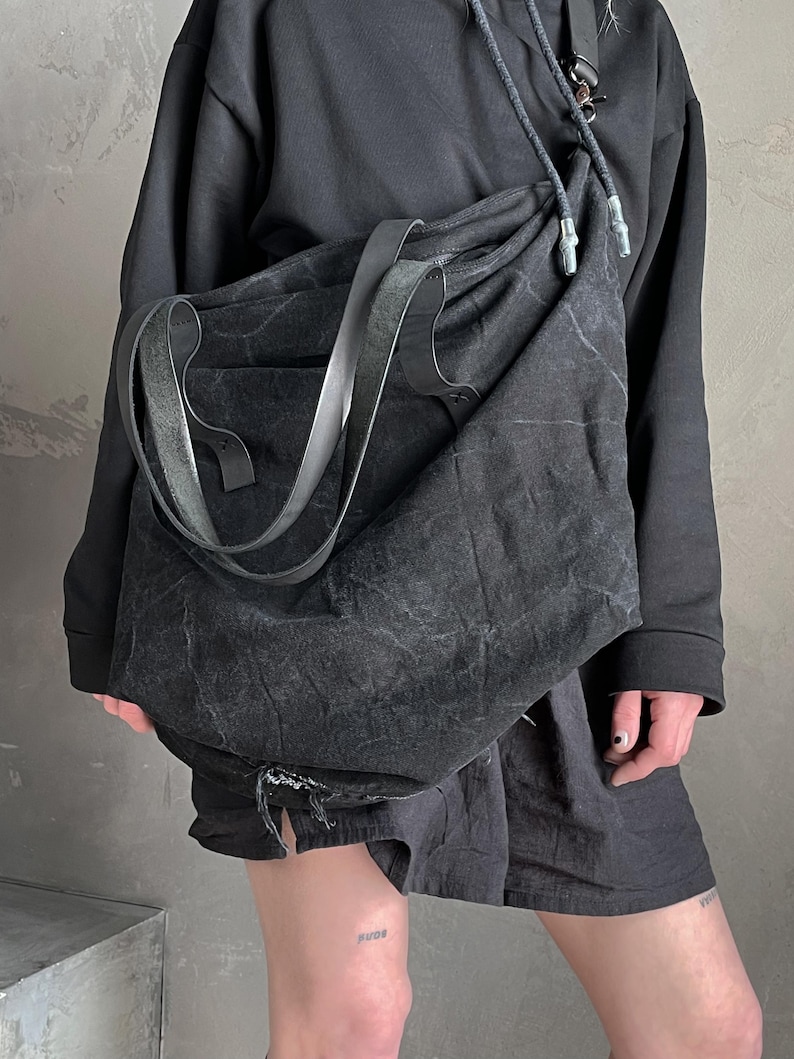May include: A large, slouchy black shoulder bag made of a textured, distressed fabric. The bag features a wide, flat strap and a drawstring closure at the top with cord ends. The material has a weathered, matte appearance with visible creases.