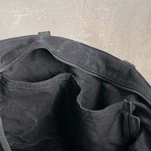 May include: The interior of a black canvas tote bag featuring a zipped main compartment closure, a single large open side pocket, and two smaller open organisational pockets along the inner lining, all constructed from matching black fabric.