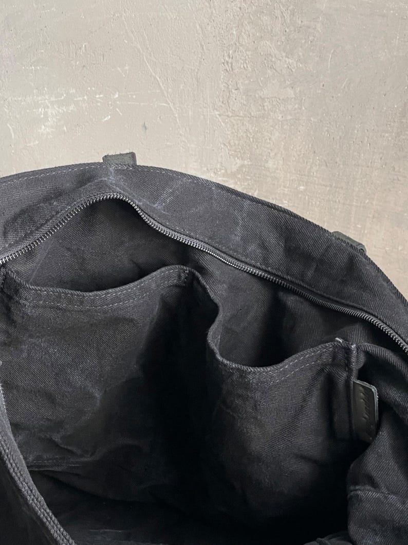 May include: The interior of a black canvas tote bag featuring a zipped main compartment closure, a single large open side pocket, and two smaller open organisational pockets along the inner lining, all constructed from matching black fabric.