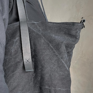 May include: A black canvas tote bag featuring a textured fabric surface and a long, smooth black leather shoulder strap. The strap is securely attached to the bag with visible cross-stitch detailing in black thread, providing a sturdy connection.