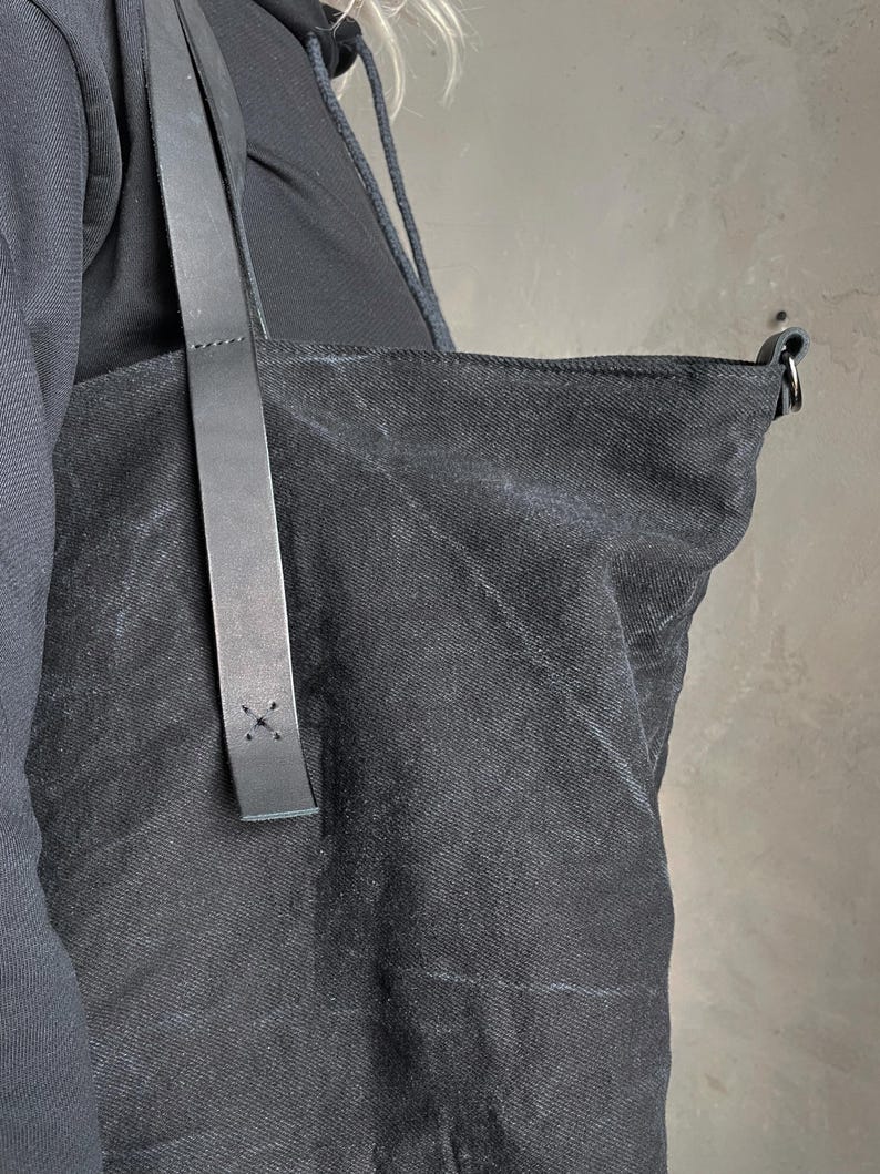 May include: A black canvas tote bag featuring a textured fabric surface and a long, smooth black leather shoulder strap. The strap is securely attached to the bag with visible cross-stitch detailing in black thread, providing a sturdy connection.