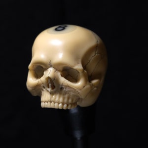 Puede incluir: Una escultura de calavera blanca con un número 8 negro en la parte superior de la cabeza. La calavera está hecha de marfil y tiene un diseño detallado.