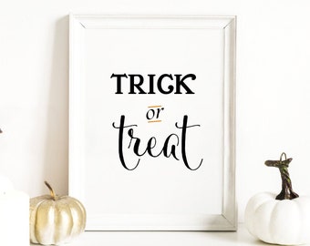 Puede incluir: Una impresión enmarcada en blanco con el texto "TRICK or treat" en letras negras. La impresión está sobre una superficie blanca con tres calabazas blancas y una calabaza dorada.