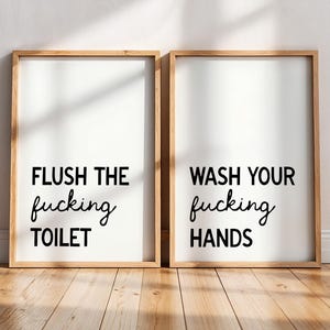 Puede incluir: Dos impresiones artísticas enmarcadas con fondos blancos y marcos de madera. La impresión izquierda dice "FLUSH THE fucking TOILET" y la derecha "WASH YOUR fucking HANDS" en escritura negra. Las impresiones se muestran en un suelo de madera.