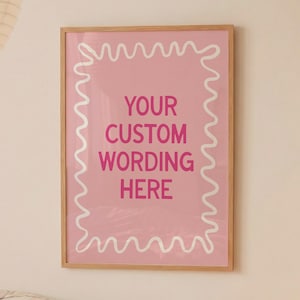 Puede incluir: Un póster rosa enmarcado con el texto "YOUR CUSTOM WORDING HERE" en una fuente lúdica. El póster tiene un borde ondulado blanco y un marco de madera. El póster está colgado en una pared blanca.