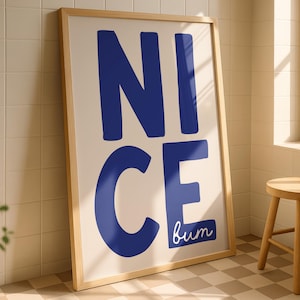 May include: A framed art print with the words "NICE" and "bum" in blue on a cream background. The print is in a light wood frame and leans against a tiled wall. A small wooden stool sits nearby.