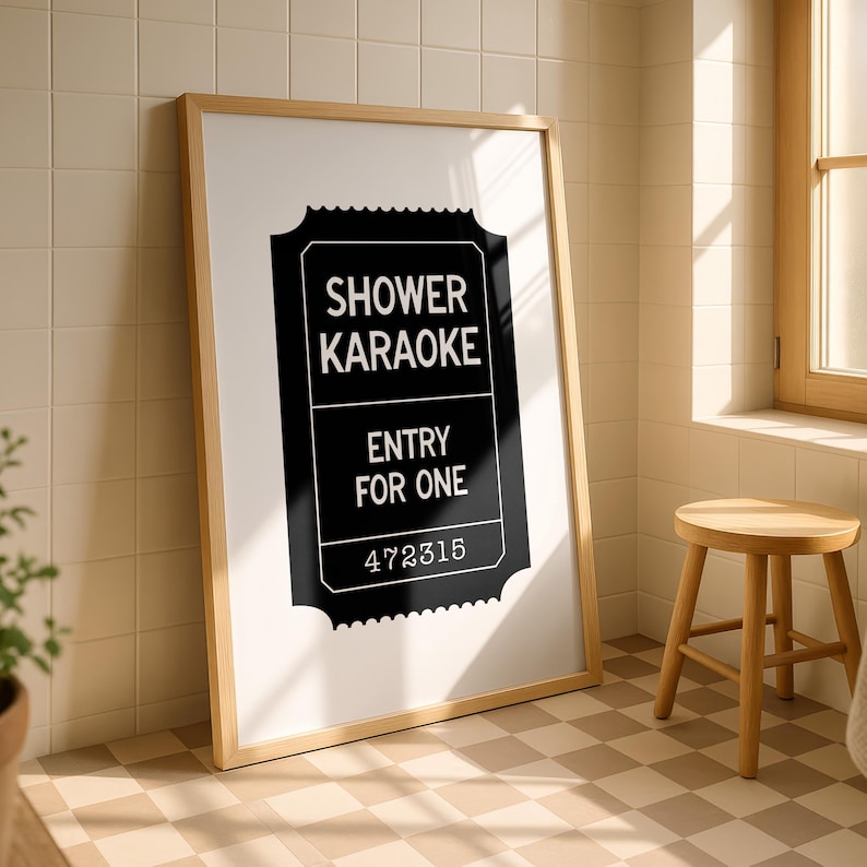 May include: A framed print with the text "SHOWER KARAOKE ENTRY FOR ONE" in a black ticket design. The print is in a light wood frame, leaning against a tiled wall. A small wooden stool sits nearby.