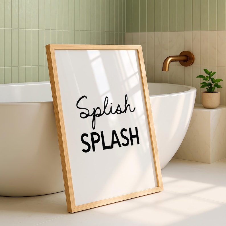 May include: A framed print with the words "Splish SPLASH" in black script and block letters. The frame is light wood, and the background is white. The print is leaning against a white bathtub in a bathroom setting.