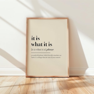 May include: A framed print with the phrase "it is what it is" in black text on a cream background. The frame is a light brown color, and the print is leaning against a white wall on a wooden floor.