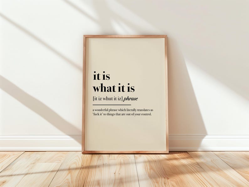 May include: A framed print with the phrase "it is what it is" in black text on a cream background. The frame is a light brown color, and the print is leaning against a white wall on a wooden floor.