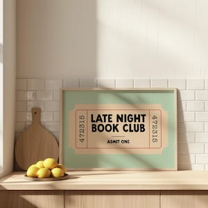 May include: A framed print with a light green background and a ticket design that reads "LATE NIGHT BOOK CLUB ADMIT ONE." The print is in a light wood frame and sits on a wooden surface. A wooden cutting board and a plate of lemons are also visible.