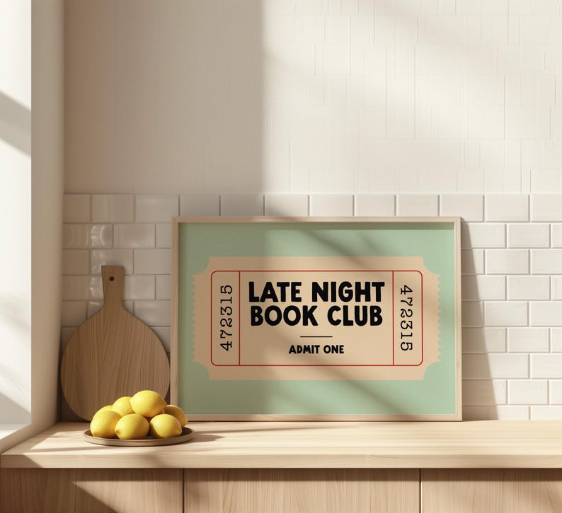 May include: A framed print with a light green background and a ticket design that reads "LATE NIGHT BOOK CLUB ADMIT ONE." The print is in a light wood frame and sits on a wooden surface. A wooden cutting board and a plate of lemons are also visible.