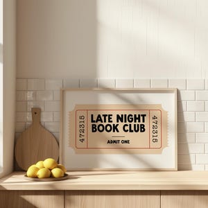 May include: A framed print with the text "LATE NIGHT BOOK CLUB" in black on a tan ticket design. The print is displayed on a wooden surface with a cutting board and a plate of lemons.