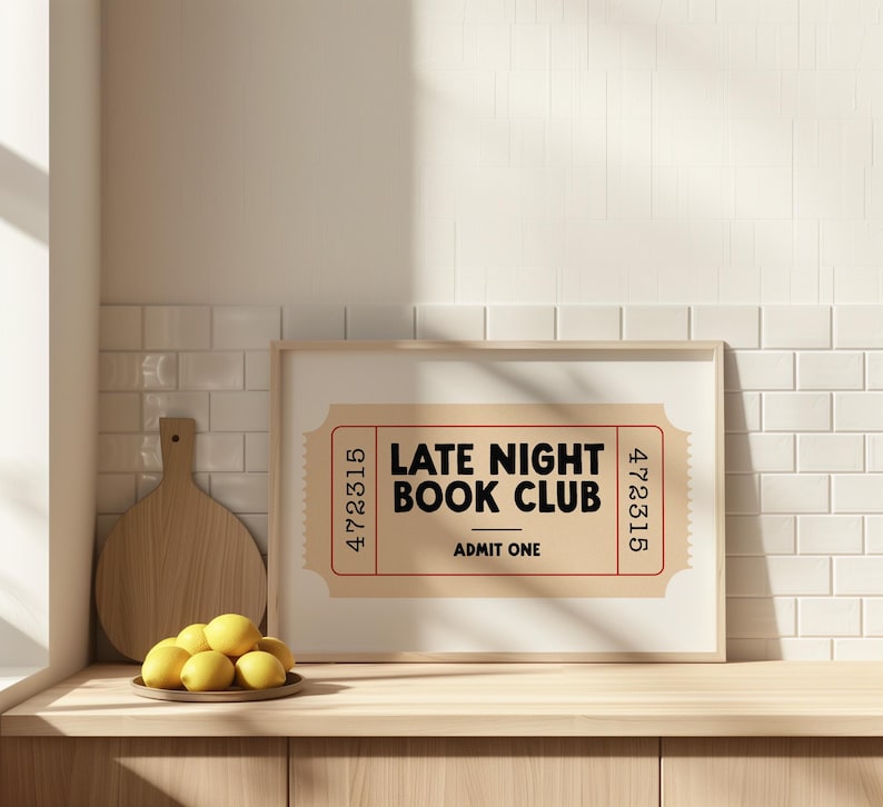 May include: A framed print with the text "LATE NIGHT BOOK CLUB" in black on a tan ticket design. The print is displayed on a wooden surface with a cutting board and a plate of lemons.