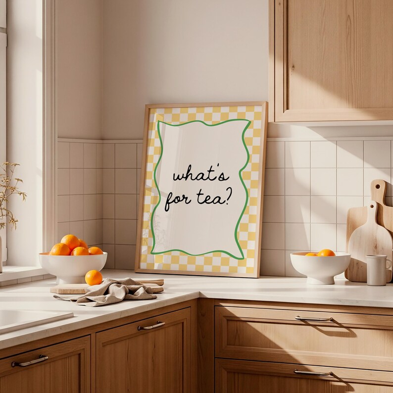 Peut inclure: Une affiche encadr&eacute;e avec le texte "what's for tea ?" dans une cuisine. L'affiche a une bordure &agrave; carreaux jaunes et blancs avec un contour vert ondul&eacute;. Un bol d'oranges est pos&eacute; sur le comptoir.