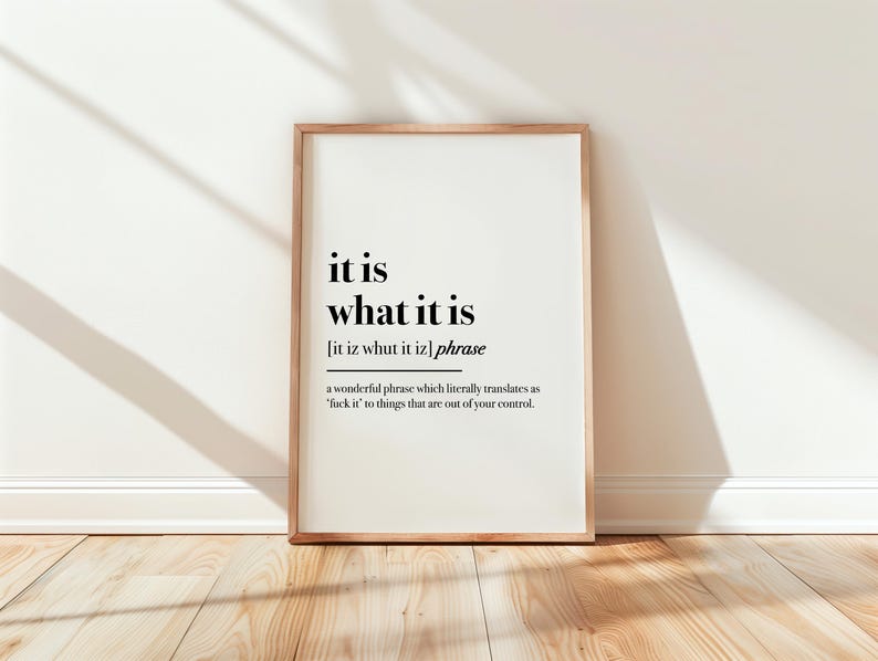 May include: A framed print with the phrase "it is what it is" in black text on a white background. The frame is a light brown color. The print is leaning against a white wall on a wooden floor.