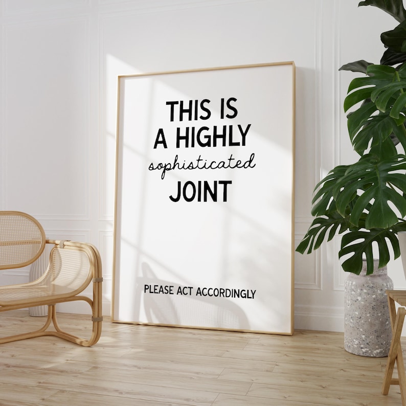 Op de afbeelding: Een ingelijste kunstdruk met de tekst "THIS IS A HIGHLY sophisticated JOINT PLEASE ACT ACCORDINGLY" in zwarte letters. De print heeft een gouden lijst en leunt tegen een witte muur. Een rieten stoel en een plant in pot staan op de achtergrond.
