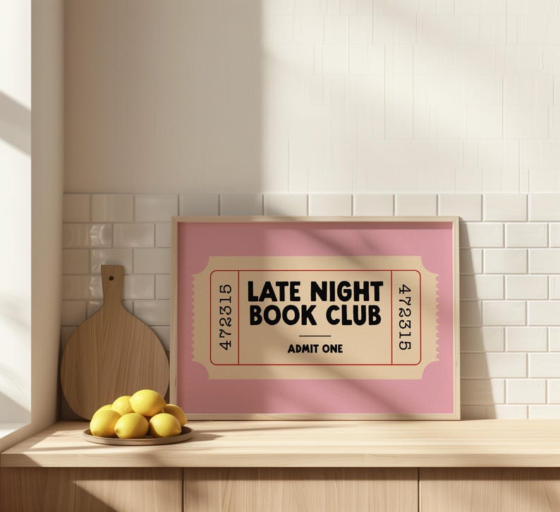 May include: A framed pink print with the text "LATE NIGHT BOOK CLUB" in black, designed like a vintage ticket. A wooden cutting board and a plate of lemons sit nearby on a wooden surface.