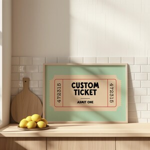 May include: A framed print featuring a vintage-style ticket design with the words "CUSTOM TICKET" and "ADMIT ONE" in bold black text. The ticket has a light brown background with red border and numbers. The frame is light brown.