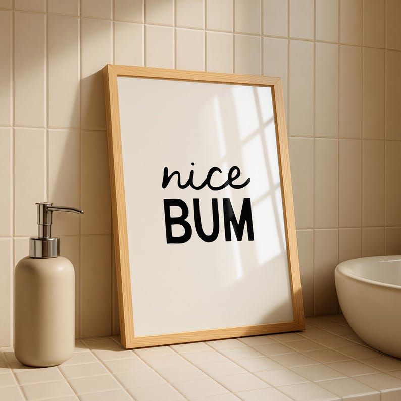 May include: A framed print with the words "nice BUM" in black script and block letters. The frame is light wood. A beige soap dispenser with a silver pump is to the left of the frame. The background is a tiled wall.