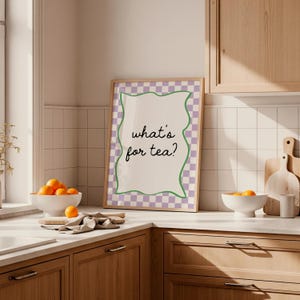 Peut inclure: Une affiche encadr&eacute;e avec le texte "what's for tea ?" dans une police fantaisiste. L'affiche a un fond blanc, une bordure verte ondul&eacute;e et un motif &agrave; carreaux violets et blancs. Le cadre est de couleur marron clair.