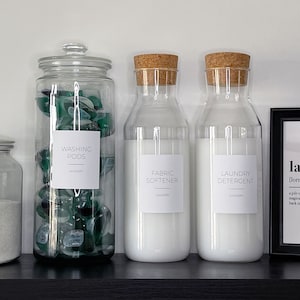 May include: Four glass jars with labels on a black shelf. The jars contain laundry detergent, fabric softener, washing pods, and oxy powder. A framed print with the word "laundry" and its definition is on the shelf.