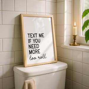 May include: A framed print with the text "TEXT ME IF YOU NEED MORE loo roll" in black on a white background. The frame is light wood. The print rests on a white toilet, with a beige towel hanging below. A lit candle sits on a windowsill.