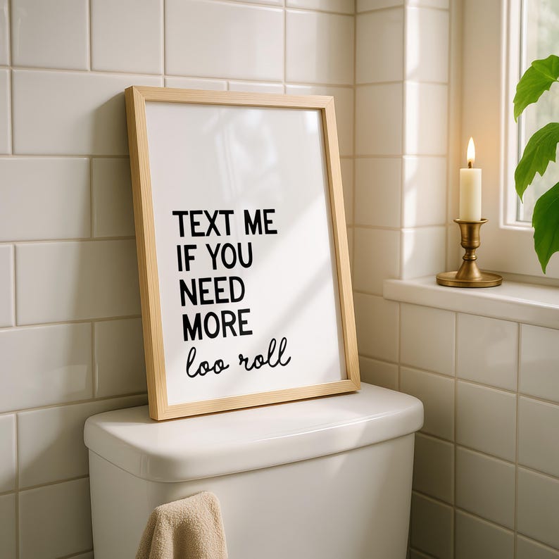 May include: A framed print with the text "TEXT ME IF YOU NEED MORE loo roll" in black on a white background. The frame is light wood. The print rests on a white toilet, with a beige towel hanging below. A lit candle sits on a windowsill.