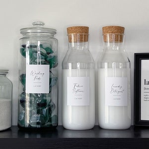 May include: Four glass jars with labels for laundry supplies: washing pods, fabric softener, laundry detergent, and a jar of powder. The jars are on a black shelf with a framed print that says "laundry" and its definition.