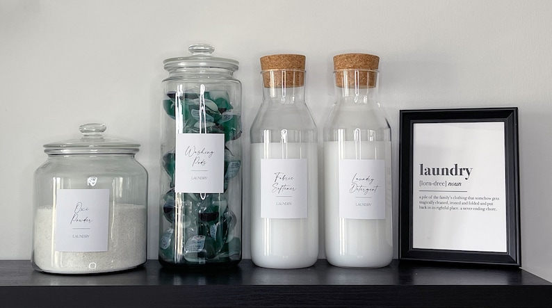 May include: Four glass jars with labels for laundry supplies: washing pods, fabric softener, laundry detergent, and a jar of powder. The jars are on a black shelf with a framed print that says "laundry" and its definition.