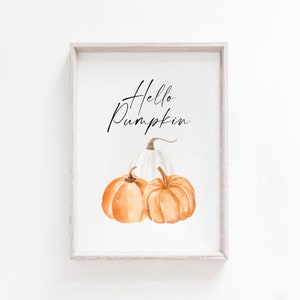 May include: A framed watercolour print featuring three pumpkins. Two orange pumpkins are in front of a white pumpkin. The words "Hello Pumpkin" are written in black script above the pumpkins. The frame is a light grey colour.
