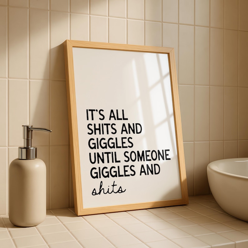 May include: A framed print with the text "IT'S ALL SHITS AND GIGGLES UNTIL SOMEONE GIGGLES AND shits" in black lettering. A beige soap dispenser with a silver pump is to the left of the frame. The background is a tiled wall.