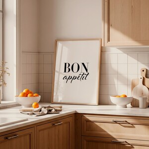 May include: A framed print with the words "BON app&eacute;tit" in black script. The kitchen scene includes a white bowl of oranges, a wooden cutting board, and a white bowl with oranges. The kitchen has wooden cabinets and white tile.