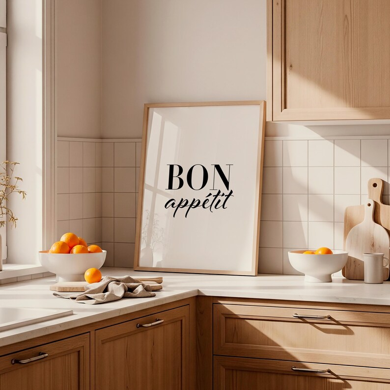 May include: A framed print with the words "BON app&eacute;tit" in black script. The kitchen scene includes a white bowl of oranges, a wooden cutting board, and a white bowl with oranges. The kitchen has wooden cabinets and white tile.