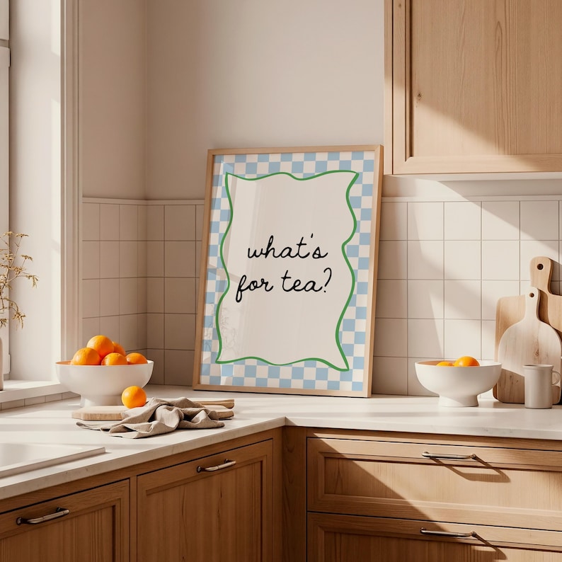 Peut inclure: Une affiche encadr&eacute;e avec le texte "what's for tea?" dans une police fantaisiste. L'affiche a une bordure &agrave; carreaux bleus et blancs avec un contour vert ondul&eacute;. L'image est dans une cuisine avec des oranges dans un bol blanc.