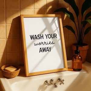 May include: A framed print with the words "WASH YOUR worries AWAY" in black script. The wooden frame sits on a bathroom sink next to a soap dispenser, a small woven basket with soap, and a potted plant.