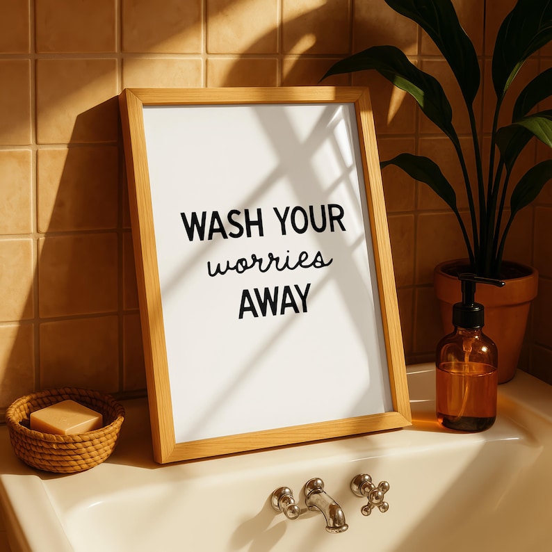 May include: A framed print with the words "WASH YOUR worries AWAY" in black script. The wooden frame sits on a bathroom sink next to a soap dispenser, a small woven basket with soap, and a potted plant.