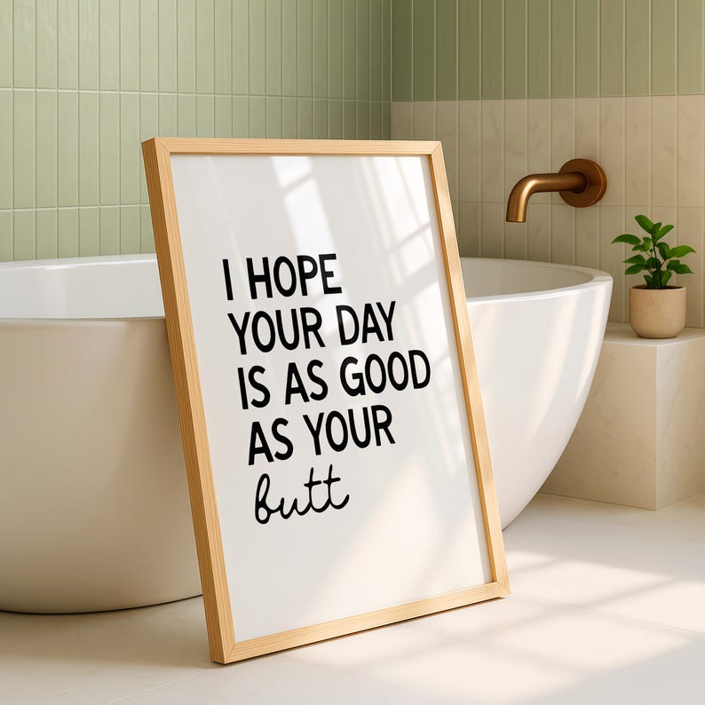 May include: A framed print with the text "I HOPE YOUR DAY IS AS GOOD AS YOUR butt" in black font. The frame is light brown. The print is leaning against a white bathtub in a bathroom setting.