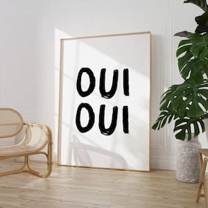 May include: A black and white art print with the word "OUI" written twice in a bold, brushstroke font. The print is framed in a thin, gold frame and is hanging on a white wall.